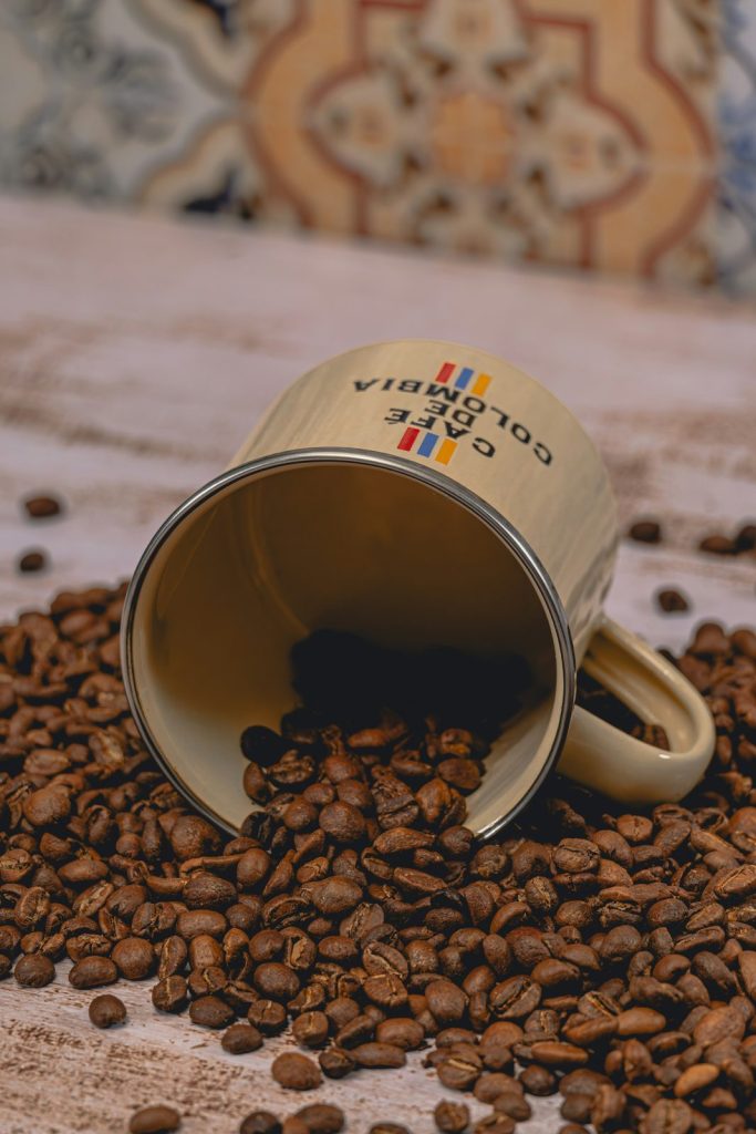 咖啡知識 印有 Café de Colombia 字樣的金色金屬杯倒在桌上,杯中灑出烘焙好的咖啡豆